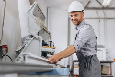 Özel üniformalı ve beyaz kasklı bir erkek işçi fabrikadaki PVC pencere ve kapı üretimi için CNC makinesinde çalışıyor..