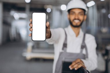 Beyaz şapkalı ve tulumlu mutlu Hindu işçilerin portresi akıllı telefon tutuyor ve fabrika arka planında boş ekran gösteriyor..