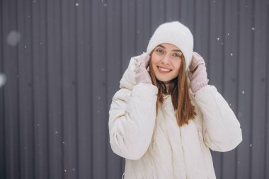Örgü eldivenli mutlu bir kadın, kışın kar yağarken gri bir duvarın arkasında poz verir.
