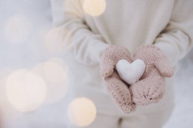 Örgü eldivenler takan kadın elleri, kışın arka planda bir kardan kalp tutar.