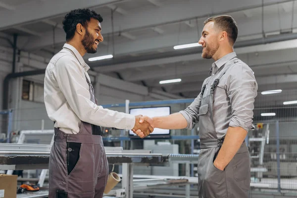 Two international male workers in overalls shake hands against the background of the production of PVC windows and doors
