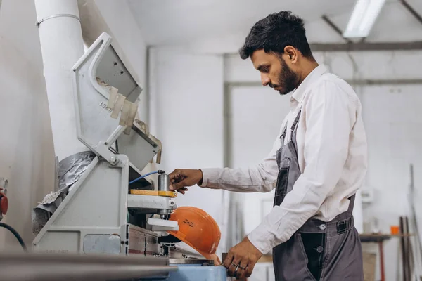 A male Indian worker in a special uniform and an orange helmet works on a CNC machine for the production of PVC windows and doors at the factory.