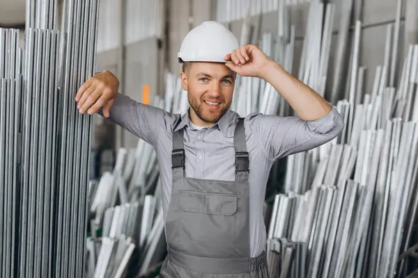 金属のプラスチック窓およびドアの生産でアルミニウム フレームを保持する特別な制服および白い堅い帽子の幸せなひげを付けられた男性の工場労働者 — ストック写真