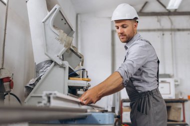 Özel üniformalı ve beyaz kasklı bir erkek işçi fabrikadaki PVC pencere ve kapı üretimi için CNC makinesinde çalışıyor..