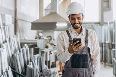 Özel üniformalı mutlu Hintli erkek fabrika işçisi ve telefonu tutan beyaz başlıklı adam metal-plastik pencere ve kapı üretiminde diyalog kuruyor.