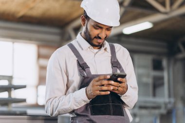 Özel üniformalı mutlu Hintli erkek fabrika işçisi ve telefonu tutan beyaz başlıklı adam metal-plastik pencere ve kapı üretiminde diyalog kuruyor.