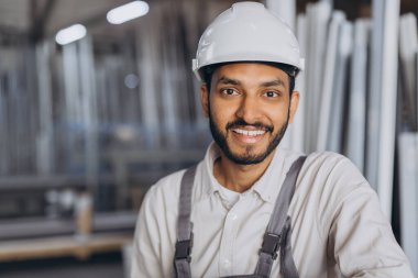 Beyaz kasketli ve tulumlu mutlu bir Hindu işçinin portresi bir fabrika ve alüminyum çerçevelerin arka planına karşı hidrolik bir kamyon tutuyor..