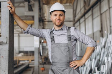 Gri üniformalı mutlu bir işçinin portresi ve fabrika yapımının arka planında poz veren beyaz bir şapka.