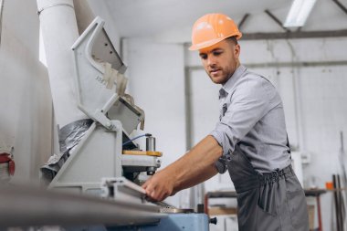 Özel üniformalı ve turuncu kasklı bir erkek işçi fabrikadaki PVC pencere ve kapı üretimi için CNC makinesinde çalışıyor..