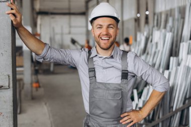 Gri üniformalı mutlu bir işçinin portresi ve fabrika yapımının arka planında poz veren beyaz bir şapka.