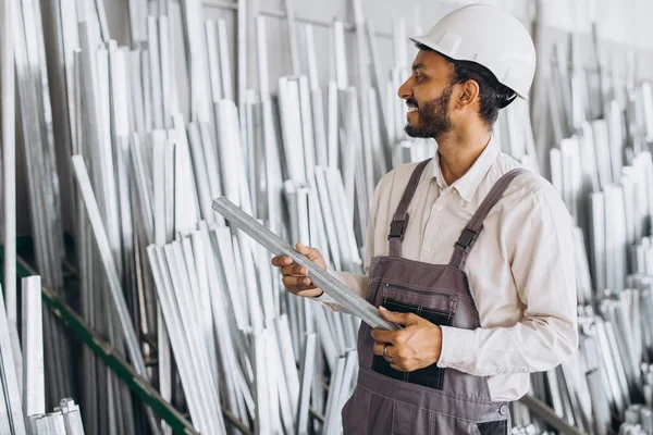 Happy bearded Indian male factory worker in special uniform and white hard hat holding aluminum frame at production of metal plastic windows and doors