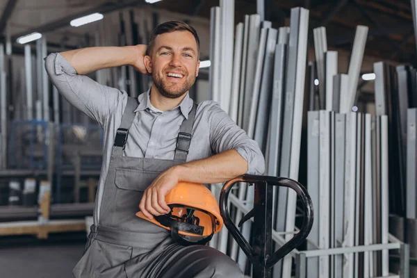 Portrait of a happy worker in a orange hard hat and overalls holding a hydraulic truck against a background of a factory and aluminum frames.