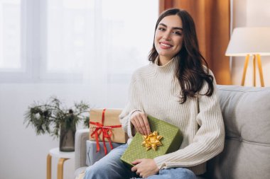 Güzel, mutlu bir kadın yeni yıl paketini açıyor ya da Noel hediyelerini kanepeye koyuyor.