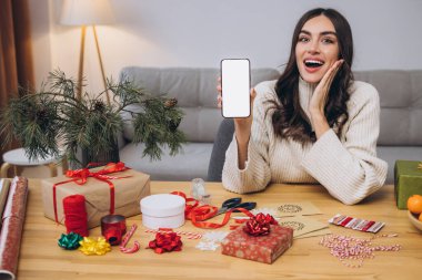 Güzel, mutlu bir kadın yılbaşı ya da Noel hediyesi paketlerken elinde fotokopi alanı olan boş ekranlı akıllı telefon arkaplanı tutarken ya da evinde taklit yaparken.