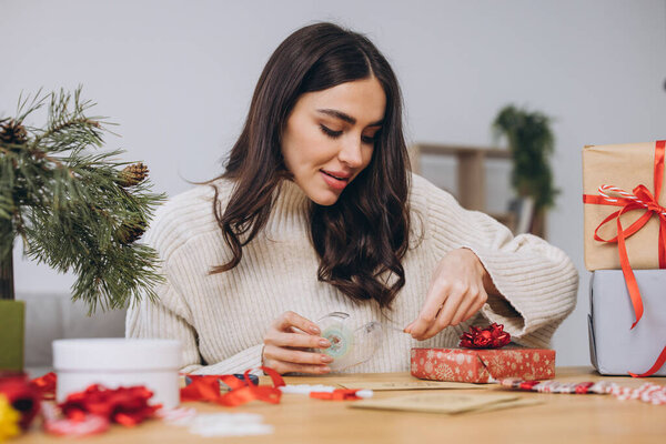 Женщина, упаковывающая рождественские подарочные коробки, закрывай. Неподготовленные подарки на столе с элементами декора и элементами рождественской или новогодней упаковки.