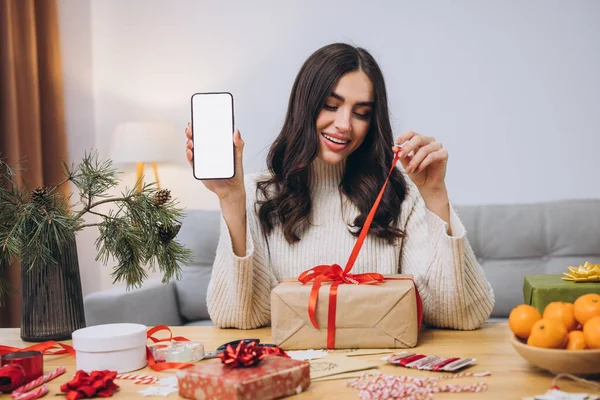 Güzel, mutlu bir kadın yılbaşı ya da Noel hediyesi paketlerken elinde fotokopi alanı olan boş ekranlı akıllı telefon arkaplanı tutarken ya da evinde taklit yaparken.