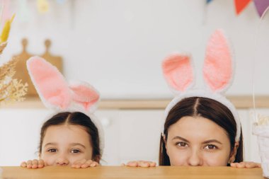 Mutlu Paskalyalar! Genç anne ve tatlı kız masanın arkasında saklanırken tavşan kulağı takıyor ve evde mutfakta biraz eğleniyorlar.