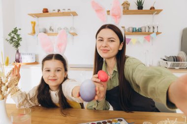 Anne ve kızı yumurta boyuyor ve selfie ya da video görüşmesi yapıyorlar. Mutlu aile Paskalya için hazırlanıyor. Tavşan kulağı takan şirin bir kız çocuğu..