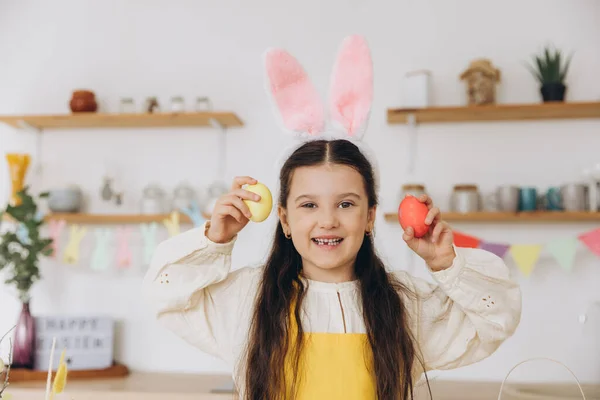 复活节快乐 可爱的小女孩手里拿着五彩缤纷的复活节彩蛋 在家里的厨房里玩得很开心 复活节那天 小女孩戴着小兔子耳朵 — 图库照片