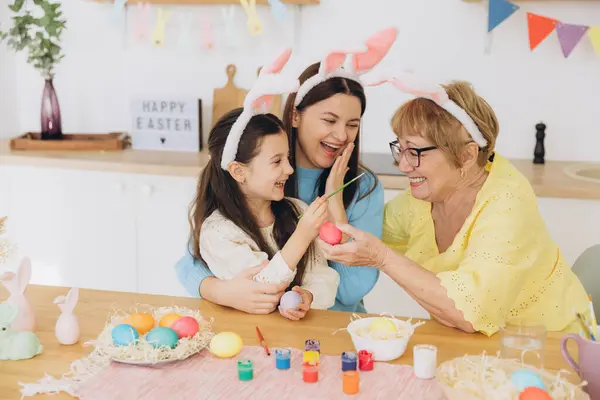 Happy Easter! Three generations of women, happy mother with daughter and grandmother painting colorful eggs and having a good time together in the kitchen