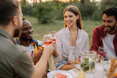 Çok ırklı bir grup mutlu arkadaş masanın etrafında oturur, şarap içer ve yaz ormanlarında akşam yemeği partisinde konuşurlar.