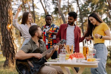 Çok kültürlü bir grup insan, sakallı hippi gitar çalıyor, arkadaşları dans ediyor ve eğleniyor.