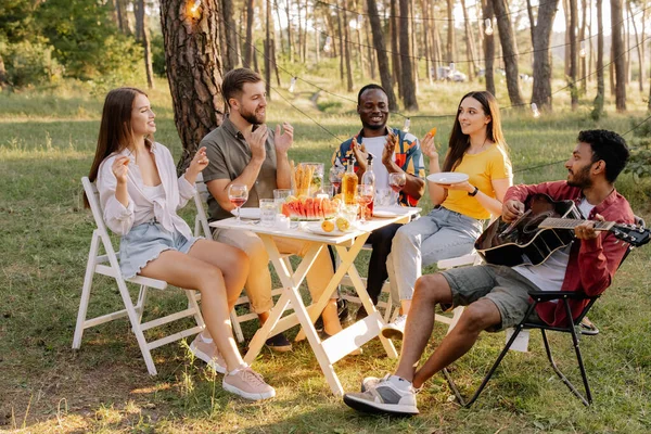 Meeting of multiracial group of friends playing guitar, singing, eating dinner and drinking wine during party in the forest