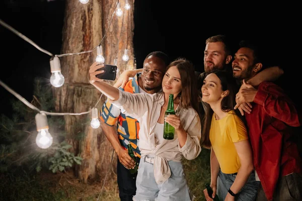 一群多种族的朋友晚上在吊灯前自娱自乐 喝啤酒 — 图库照片