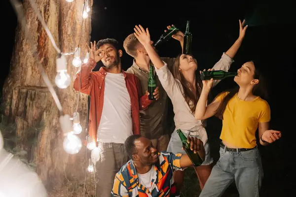 A company of multiracial friends drinking beer at party, making faces near hanging lamps