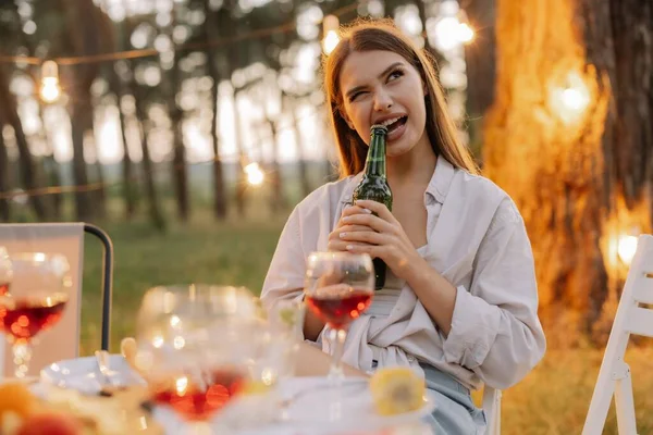 Funny hipster woman opens beer with teeth at party with friends in forest decorated with hanging lamps