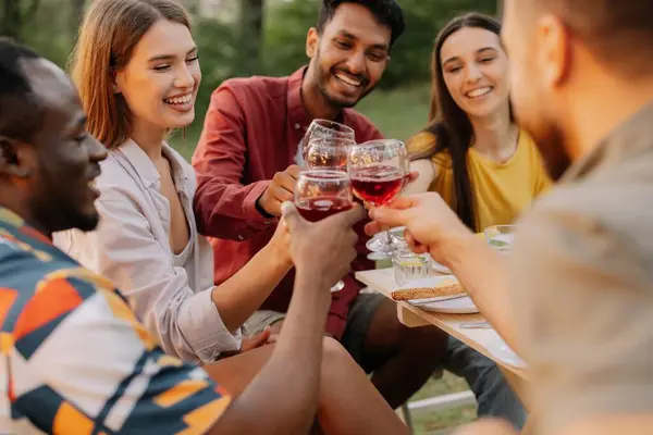 Multiracial group of happy friends sitting around table drinking wine talking at dinner party in evening summer forest