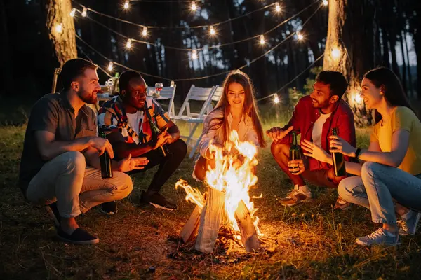 Multiracial friends sitting around the fire drinking beer and having fun at the party
