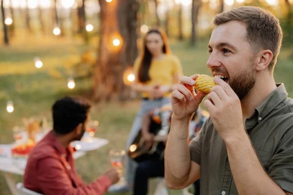 Bearded hipster man eating corn on the background of dinner party of multiracial friends