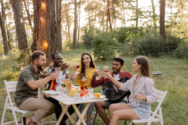 Meeting of multiracial group of friends playing guitar, singing, eating dinner and drinking wine during party in the forest