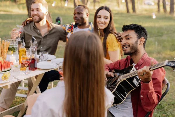 Multiracial group of people, Indian hipster man playing guitar and friends eating dinner and drinking wine during party in the forest