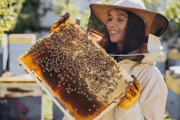 Happy smiling female Beekeeper in protective suit holding honeybee frame with bees at apiary