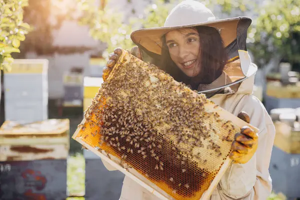 Happy smiling female Beekeeper in protective suit holding honeybee frame with bees at apiary