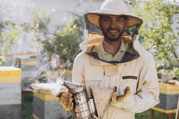Happy smiling Indian male Beekeeper smoking honey bees with bee smoker on the apiary