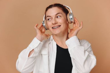 Dişlerinde diş telleriyle gülümseyen ve bej arka planda kulaklıkla müzik dinleyen modern bir kadın.