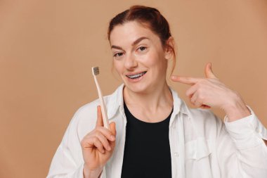 Diş teli takmış, bej arka planda diş fırçası tutan mutlu olgun bir kadın.