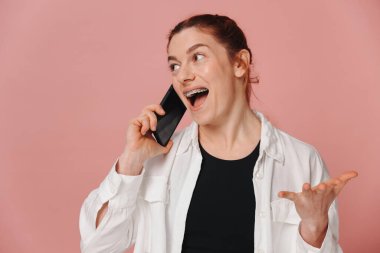 Modern mutlu kadın tellerle gülümsüyor ve pembe arka planda akıllı telefondan konuşuyor.