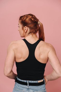 Tişörtlü ve kot pantolonlu kaslı kadın pembe arka planda güçlü görünüyor.