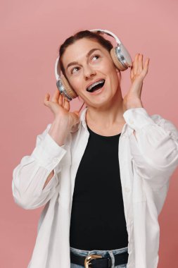 Dişlerinde diş telleriyle gülümseyen ve pembe arka planda kulaklıkla müzik dinleyen modern bir kadın.