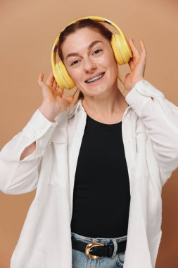 Dişlerinde diş telleriyle gülümseyen ve bej arka planda kulaklıkla müzik dinleyen modern bir kadın.
