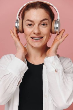 Dişlerinde diş telleriyle gülümseyen ve pembe arka planda kulaklıkla müzik dinleyen modern bir kadın.