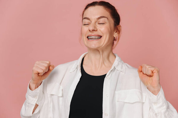 Modern happy woman smiling with braces clenching fists showing win on pink background