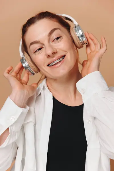 stock image Modern woman smiling with braces on her teeth and listening to music in headphones on a beige background