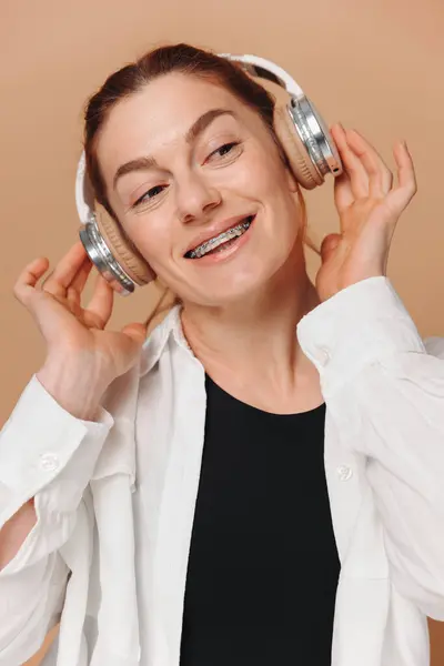 Nowoczesna Kobieta Uśmiechnięta Aparatem Zębach Słuchająca Muzyki Słuchawkach Beżowym Tle Obraz Stockowy