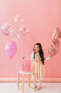 Pembe elbiseli sevimli küçük kız flamingo doğum günü pastasının yanında poz veriyor ve pembe arka planda beşinci doğum gününü kutluyor.