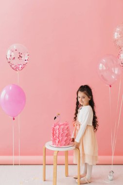 Pembe elbiseli sevimli küçük kız flamingo doğum günü pastasının yanında poz veriyor ve pembe arka planda beşinci doğum gününü kutluyor.